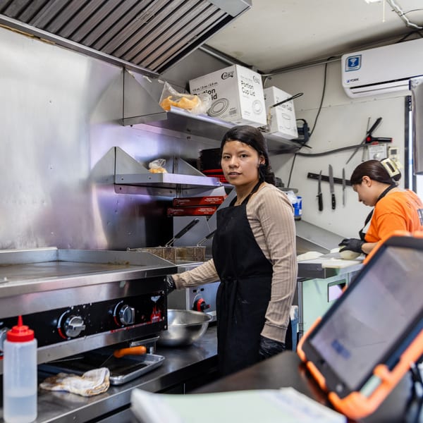 A woman in a black apron stands by a griddle in a busy El Chapin kitchen.