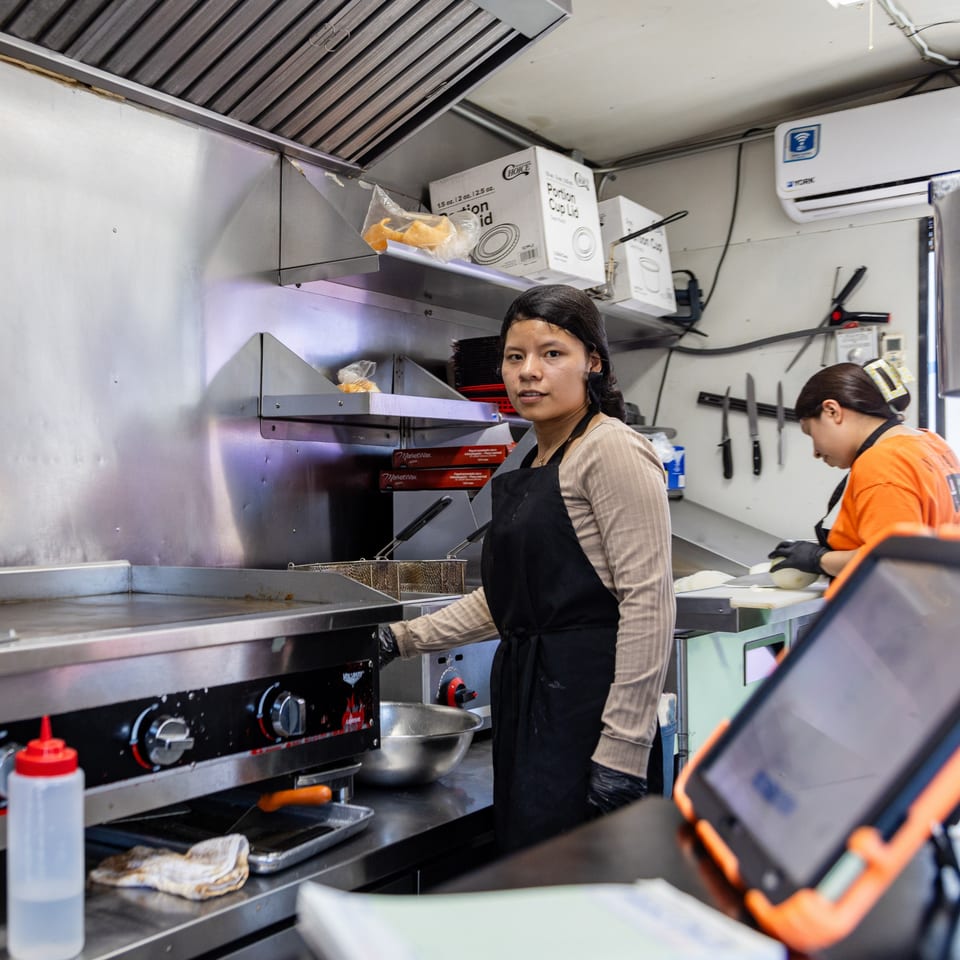 A woman in a black apron stands by a griddle in a busy El Chapin kitchen.