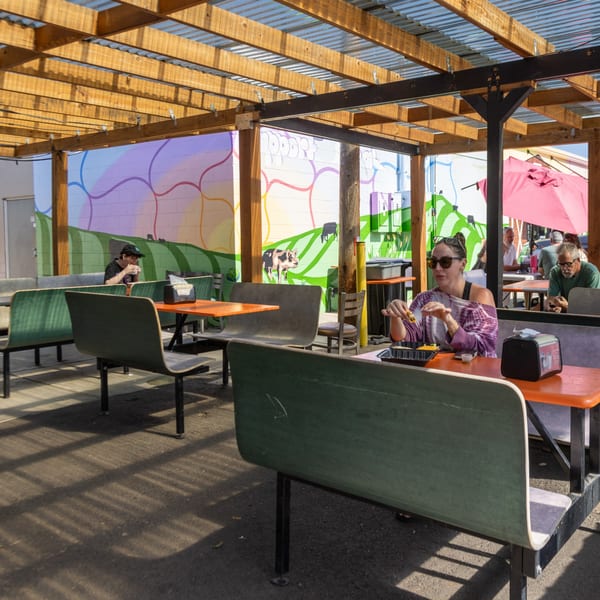 Outdoor seating area with people eating at orange tables and green benches, under a wooden pergola.