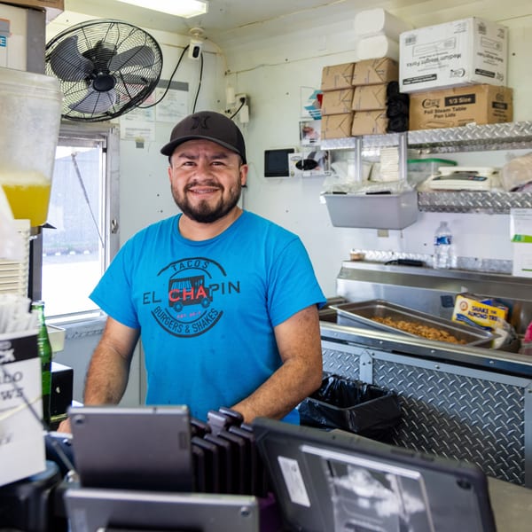 Smiling man in an "El Chapin" t-shirt stands inside a food truck kitchen.