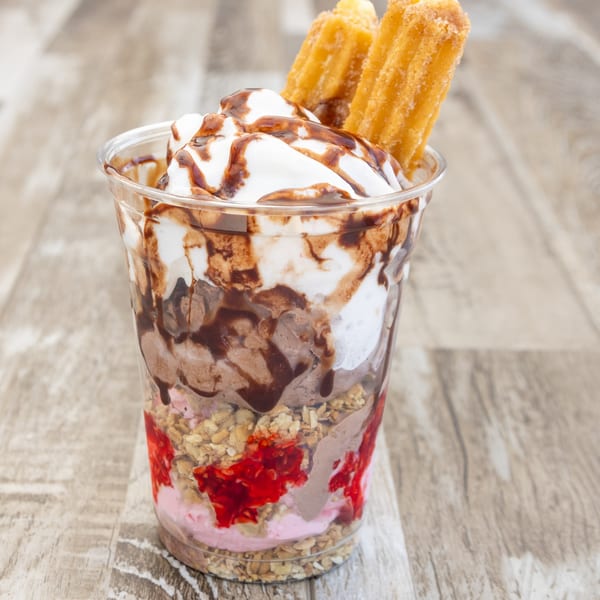A clear cup filled with layers of ice cream, granola, strawberry, whipped cream, chocolate syrup, and churros.