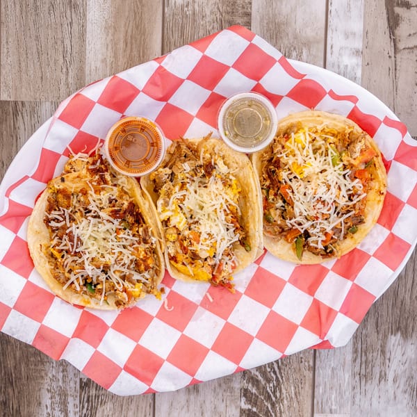 Three El Chapin breakfast tacos with shredded cheese and two sauce cups on a red and white checkered liner.