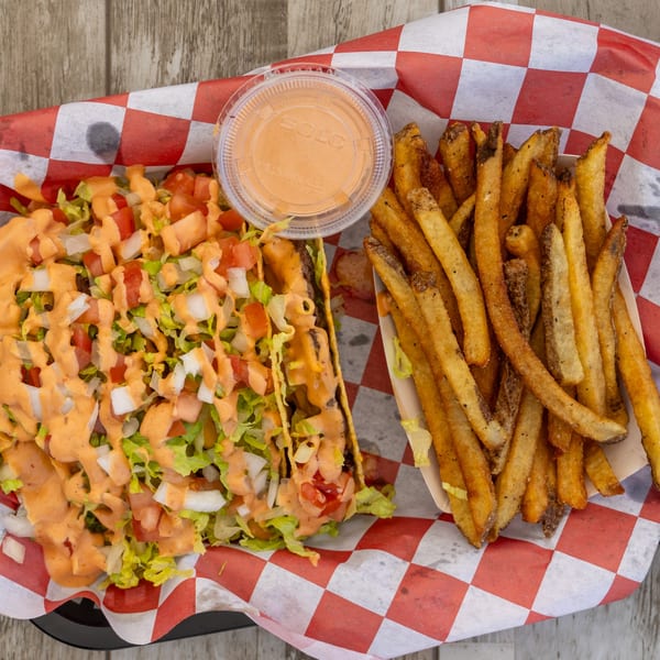 Two El Chapin tacos with lettuce, tomato, onion, and sauce, alongside a container of fries and dipping sauce.