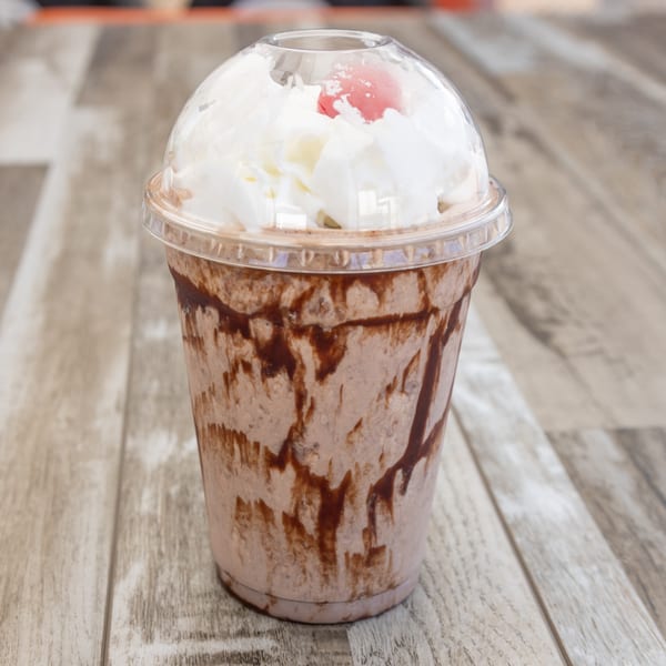 Chocolate milkshake with whipped cream and a cherry on top, served in a clear plastic cup.