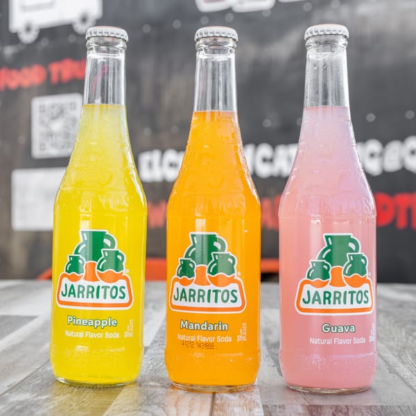 Three Jarritos sodas in pineapple, mandarin, and guava flavors on a wooden surface.