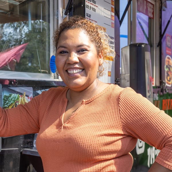 Smiling woman with curly hair in an orange shirt, standing in front of the El Chapin food truck.