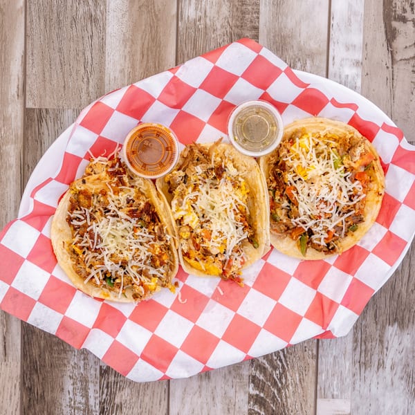 Three El Chapin breakfast tacos with shredded cheese and two sauce cups on red and white checkered paper.